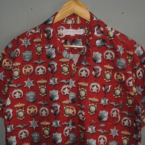 Pode incluir: Camisa vermelha de manga curta com um padr&atilde;o de distintivos de xerife, estrelas e emblemas de &aacute;guia. A camisa est&aacute; pendurada em um cabide de madeira. A marca "California Tops" &eacute; vis&iacute;vel na gola.