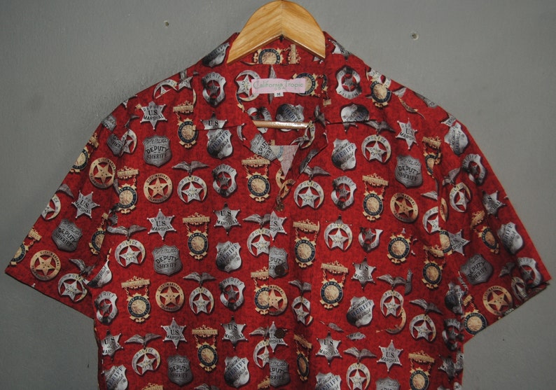 Pode incluir: Camisa vermelha de manga curta com um padr&atilde;o de distintivos de xerife, estrelas e emblemas de &aacute;guia. A camisa est&aacute; pendurada em um cabide de madeira. A marca "California Tops" &eacute; vis&iacute;vel na gola.