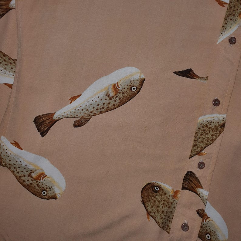 K&ouml;nnte beinhalten: Ein hellpfirsichfarbenes Hemd mit Kn&ouml;pfen, das mit detaillierten Fischillustrationen gemustert ist. Die Fische sind braun mit wei&szlig;en und beigen Akzenten, und das Hemd hat braune Kn&ouml;pfe.