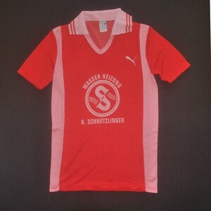 Op de afbeelding: Een rood en roze voetbaltenue met witte accenten. Het tenue heeft een Puma-logo op de rechterborst en een wit logo met de tekst "Wasser Heizung H. Schnetzlinger" op de voorkant.