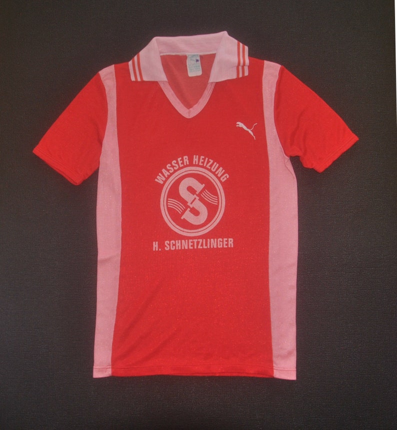 Op de afbeelding: Een rood en roze voetbaltenue met witte accenten. Het tenue heeft een Puma-logo op de rechterborst en een wit logo met de tekst "Wasser Heizung H. Schnetzlinger" op de voorkant.