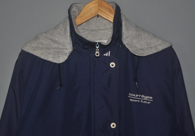 Puede incluir: Una chaqueta azul marino con capucha y forro de polar gris. La chaqueta tiene cierre de cremallera y solapa con botones. La chaqueta tiene el texto "courr&egrave;ges sport futur" en la parte delantera.