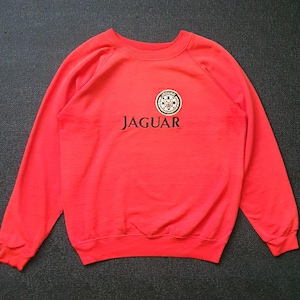 May include: A bright red long-sleeve sweatshirt featuring the word JAGUAR embroidered in black across the chest. Above the text is a circular patch with a Jaguar logo, including a stylised cat head, black and white chequered details, and text.