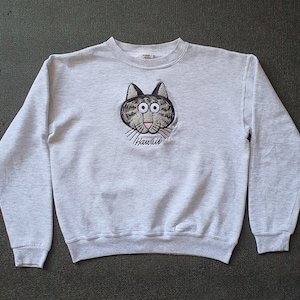 May include: A light gray sweatshirt featuring a cartoon cat face with the word "Hawaii" below. The cat has black and gray stripes, pink accents, and large eyes. The sweatshirt has long sleeves and a crew neck.