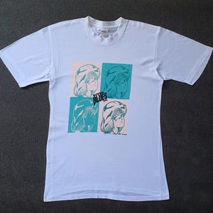 May include: White t-shirt with a graphic print of four women's faces in blue and green. The text "Stray Blues" is printed on the shirt.