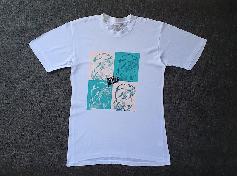 May include: White t-shirt with a graphic print of four women's faces in blue and green. The text "Stray Blues" is printed on the shirt.