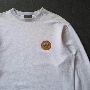 May include: A light gray sweatshirt with a crew neck and long sleeves. The sweatshirt features a small, colorful patch on the chest with a circular design and the brand name "Pendleton" on the collar.