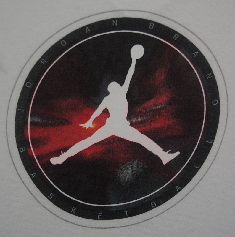 K&ouml;nnte beinhalten: Eine schwarz-wei&szlig;e Silhouette eines Basketballspielers, der mit einem Basketball in der Hand springt, in einem Kreis mit dem Text "JORDAN BRAND BASKETBALL" am Rand.