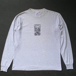 Puede incluir: Una camiseta gris de manga larga con un estampado gráfico en blanco y negro de un rostro y el texto "Kristian Jones".