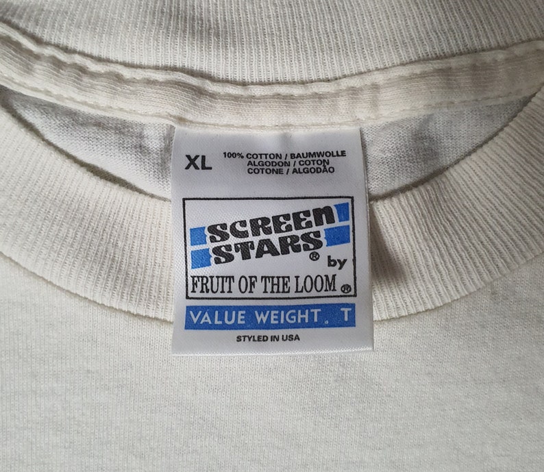 Pode incluir: Etiqueta de camiseta branca com texto azul e preto que diz "Screen Stars by Fruit of the Loom Value Weight. T Styled in USA". A etiqueta tamb&eacute;m diz "100% Algod&atilde;o/Baumwolle Algodon/Coton Cotone/Algodao" e tem uma etiqueta de tamanho que diz "XL".