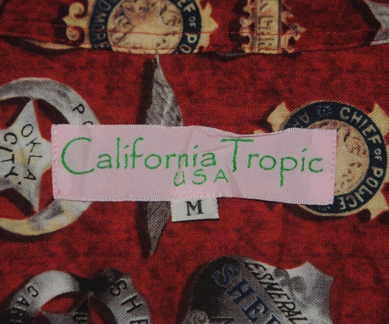 Pode incluir: Camisa vermelha estampada com distintivos e estrelas da pol&iacute;cia. A etiqueta rosa diz "California Tropic USA" e o tamanho &eacute; "M".