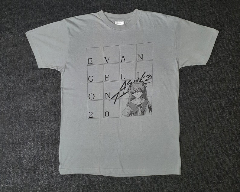 May include: A gray t-shirt with a black and white graphic print of a character from the anime series Neon Genesis Evangelion. The graphic features the text "EVANGELION" and "2.0" in a grid pattern. The character is a young woman with long, dark hair and a red and white uniform.