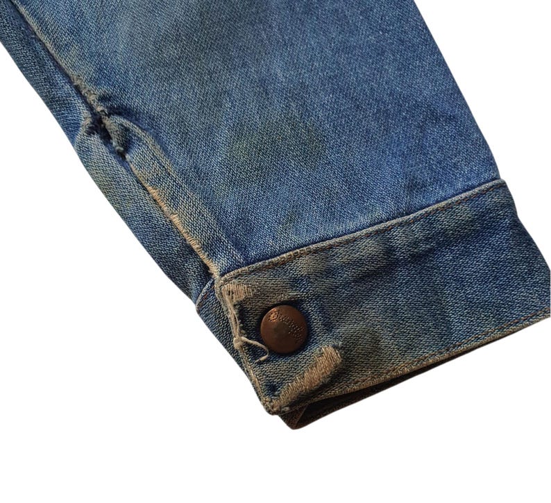 May include: Close-up of a light blue denim jacket sleeve. The cuff features a brown button and distressed edges. The denim shows signs of wear and tear, with faded areas and slight fraying.