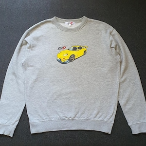 May include: A light gray crewneck sweatshirt featuring a graphic print of a yellow sports car on the chest. The graphic includes the text "Initial D" in a stylized font above the car. The garment has long sleeves, a ribbed collar, and cuffs.