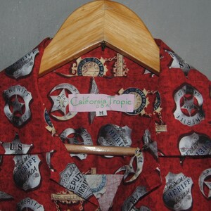 Pode incluir: Camisa vermelha com padr&atilde;o de distintivos de xerife e estrelas, pendurada em um cabide de madeira. A etiqueta diz "California Tropic USA" e o tamanho "M". A camisa tem gola e fecho de bot&otilde;es.