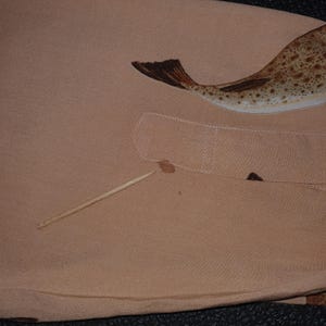 K&ouml;nnte beinhalten: Hellbraunes Stoffst&uuml;ck mit einer Illustration eines Fisches mit geflecktem K&ouml;rper und dunkelbraunem Schwanz. Ein Holz-Zahnstocher und ein kleiner Fleck sind ebenfalls auf dem Stoff zu sehen.