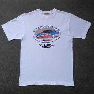 May include: White t-shirt featuring a blue and red graphic of a racing car with the text "Team Kunimitsu Racing Staff" and "VTEC Power".