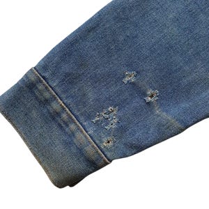 May include: Close-up of a distressed denim jacket sleeve. The blue denim shows wear and tear with several small, frayed holes near the cuff. The fabric has a faded appearance, suggesting age or a vintage style.