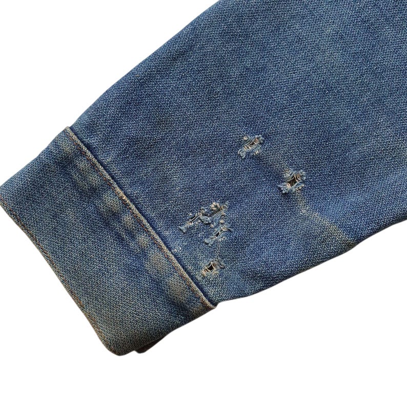 May include: Close-up of a distressed denim jacket sleeve. The blue denim shows wear and tear with several small, frayed holes near the cuff. The fabric has a faded appearance, suggesting age or a vintage style.
