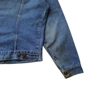 May include: Close-up of a blue denim jacket. The jacket features a classic design with a button closure and two front pockets. The denim has a slightly faded appearance, giving it a vintage look. The jacket's seams are visible, and the sleeves are long.