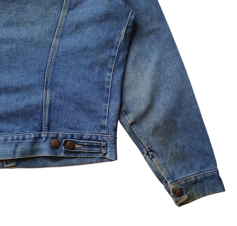 May include: Close-up of a blue denim jacket. The jacket features a classic design with a button closure and two front pockets. The denim has a slightly faded appearance, giving it a vintage look. The jacket's seams are visible, and the sleeves are long.