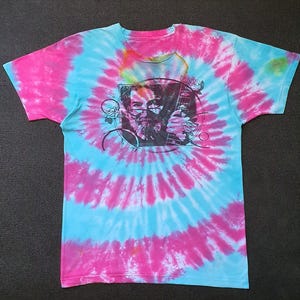 May include: Tie-dye t-shirt with a spiral pattern in pink and light blue. The shirt features a black and white graphic of a man with glasses and a guitar. The shirt is short-sleeved and made of a soft material.