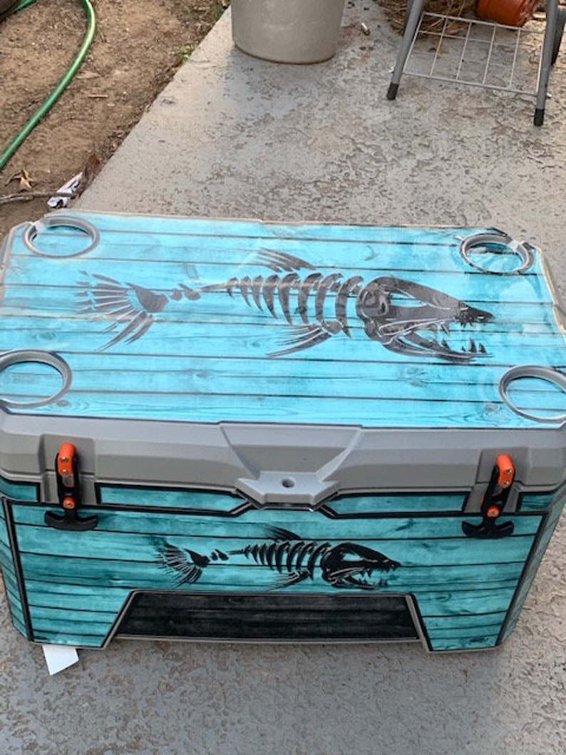 May include: A large, rectangular cooler with a teal wood-grain design and a black fish skeleton graphic. The cooler has gray accents, orange latches, and built-in cup holders. The design is repeated on the front.
