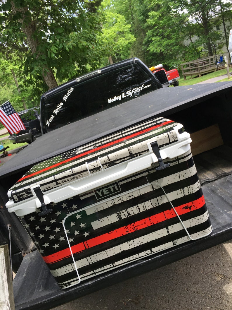 May include: A white YETI cooler with a distressed American flag design, featuring black and white stripes, stars, and a red stripe. The cooler is in the bed of a black truck.