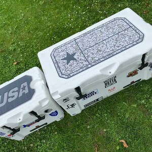 K&ouml;nnte beinhalten: Zwei wei&szlig;e K&uuml;hlboxen mit schwarzen Akzenten und verschiedenen Aufklebern. Die gr&ouml;&szlig;ere hat ein Texas-Flaggen-Design auf dem Deckel, die kleinere zeigt "USA". Beide stehen auf Gras.