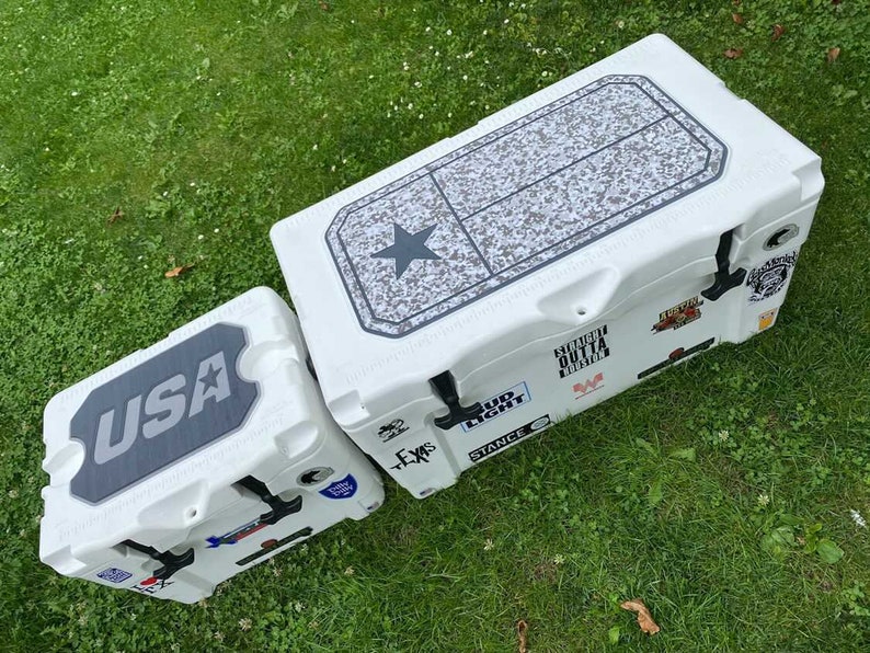 K&ouml;nnte beinhalten: Zwei wei&szlig;e K&uuml;hlboxen mit schwarzen Akzenten und verschiedenen Aufklebern. Die gr&ouml;&szlig;ere hat ein Texas-Flaggen-Design auf dem Deckel, die kleinere zeigt "USA". Beide stehen auf Gras.