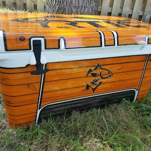 May include: A large, rectangular cooler with a wood-grain design. The cooler has a white lid and trim, black latches, and a black graphic of a fish. The cooler is sitting on grass.