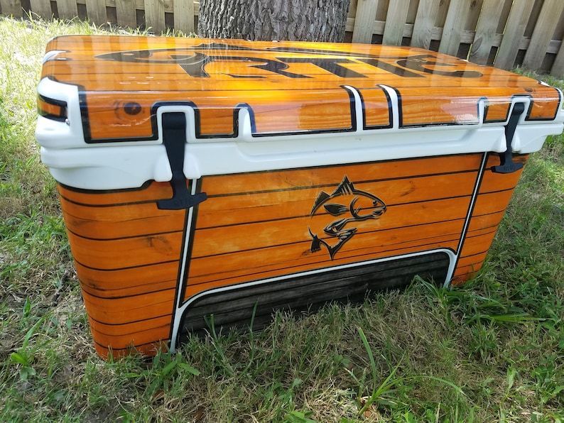 May include: A large, rectangular cooler with a wood-grain design. The cooler has a white lid and trim, black latches, and a black graphic of a fish. The cooler is sitting on grass.