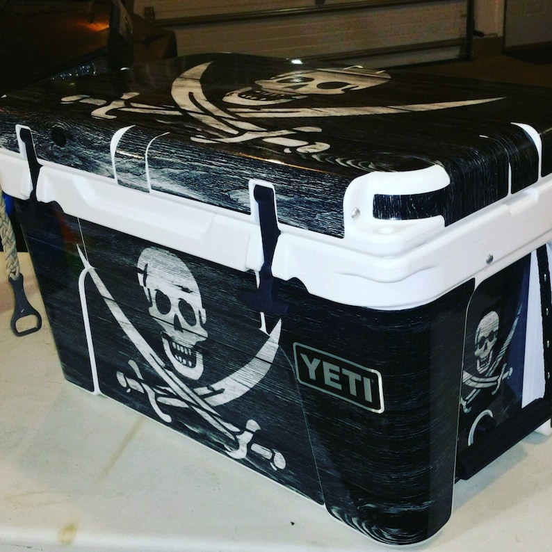 May include: A black and white cooler with a pirate flag design. The cooler features a skull and crossbones graphic, with a wood grain texture. The cooler has a white lid and trim, and the brand name "YETI" is visible.