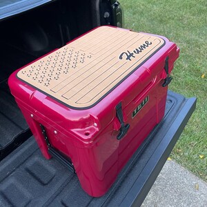 May include: A pink cooler with a wood-look lid featuring a star pattern and the name "Hume" in black lettering. The cooler is sitting in the bed of a pickup truck.