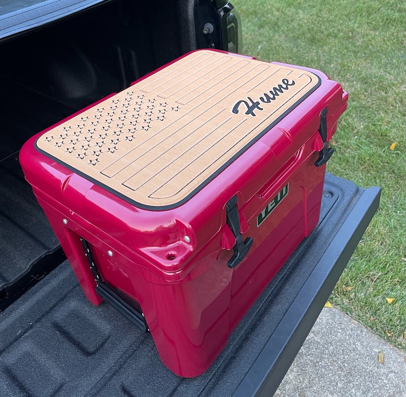 May include: A pink cooler with a wood-look lid featuring a star pattern and the name "Hume" in black lettering. The cooler is sitting in the bed of a pickup truck.