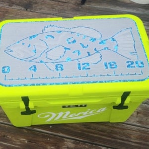 May include: A bright yellow cooler with a white "Merica" logo and a gray fish measuring ruler on the lid. The ruler is marked in inches from 0 to 20.
