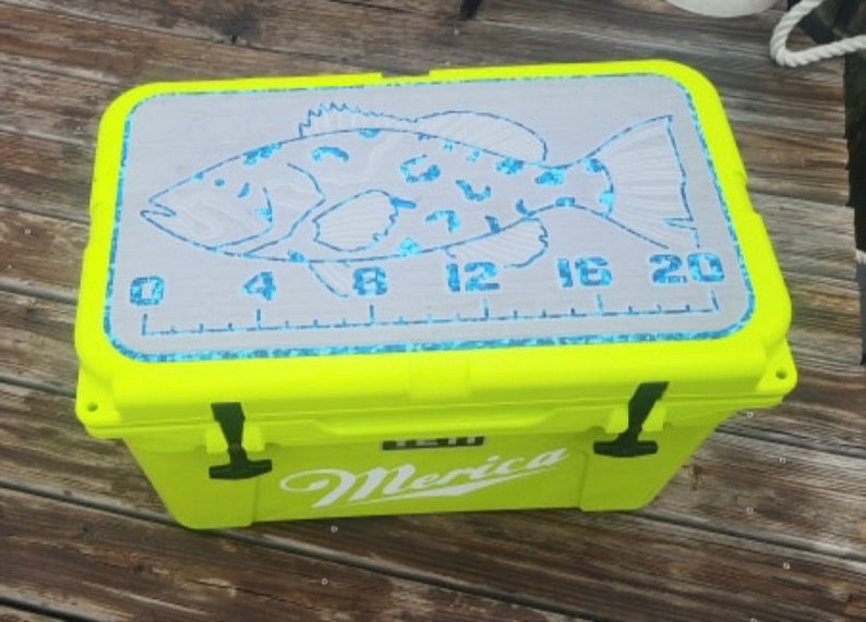 May include: A bright yellow cooler with a white "Merica" logo and a gray fish measuring ruler on the lid. The ruler is marked in inches from 0 to 20.