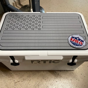 May include: White cooler with a gray American flag design on the lid. The cooler has the word "RTIC" printed on the front and a "USA Tuff-Deck" sticker on the lid.