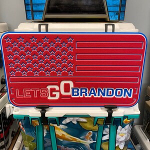 K&ouml;nnte beinhalten: Roter, wei&szlig;er und blauer K&uuml;hlboxdeckel mit amerikanischem Flaggenmuster und dem Text "LET'S GO BRANDON". Der Deckel hat einen blauen Rand und befindet sich auf einer K&uuml;hlbox mit einem Koi-Teich-Design.