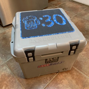 May include: A gray K2 Coolers 30-quart cooler with a black lid featuring a blue and black graphic of a beer mug and the text "3:30".