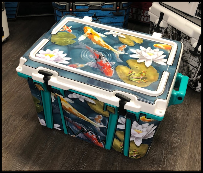 May include: A large, rectangular cooler with a teal and white exterior. The cooler lid features a detailed painting of koi fish, lily pads, and water lilies in a pond. The cooler has black latches and a teal handle.