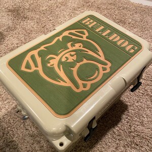 May include: A beige cooler with a green and brown bulldog graphic on the lid. The graphic says "Bulldog" in a brown font.