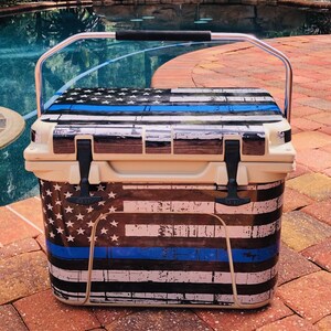 May include: A beige cooler with a distressed American flag design, featuring white stars, blue stripes, and a blue line. It has a silver handle with a black grip and black latches. The cooler is sitting on a brick surface.