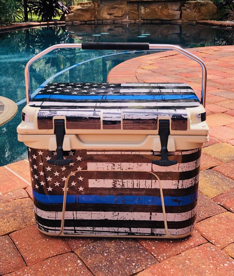 May include: A beige cooler with a distressed American flag design, featuring white stars, blue stripes, and a blue line. It has a silver handle with a black grip and black latches. The cooler is sitting on a brick surface.