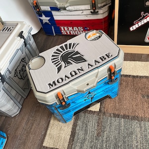 May include: A blue and gray cooler with a Spartan helmet and the text "&Mu;&Omicron;&Lambda;&Omega;&Nu; &Lambda;&Alpha;&Beta;&Epsilon;" on the lid. The cooler has a wood grain pattern and a fish design on the side.