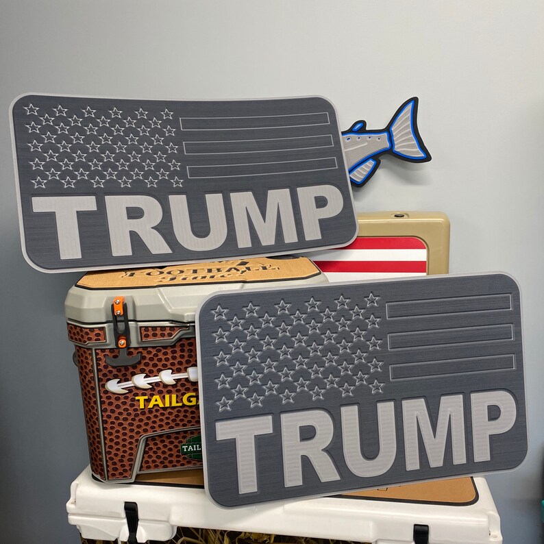 May include: Two gray rectangular signs with an American flag design and the name "TRUMP" in bold letters. The signs are displayed with a cooler featuring a football design and the word "TAILGATE". A fish-shaped decoration is also visible.