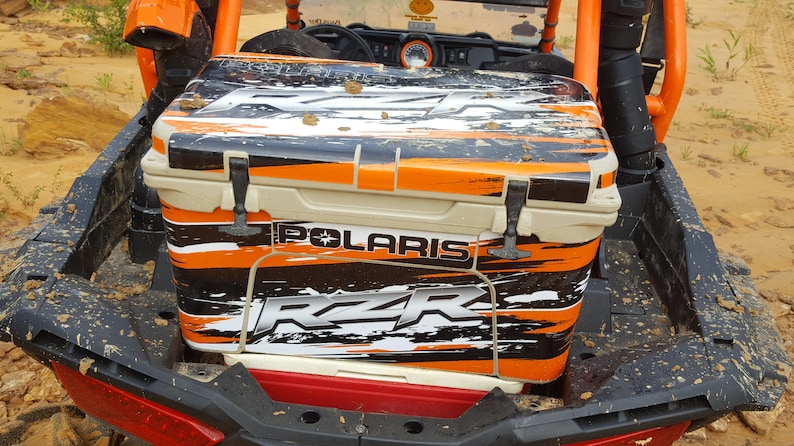 May include: A beige and black cooler with orange accents is secured in the back of an orange and black Polaris RZR. The cooler has the Polaris and RZR logos. The vehicle is on a sandy, dirt road.