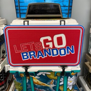 May include: Red, white, and blue cooler with a sign that says "Let's Go Brandon". The cooler has a fish and lily pad design.