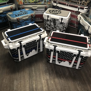 May include: Two white coolers with black and white American flag designs. One cooler has a blue stripe across the top and the other has a red stripe. Both coolers have the words "USA Tuff" on the top.