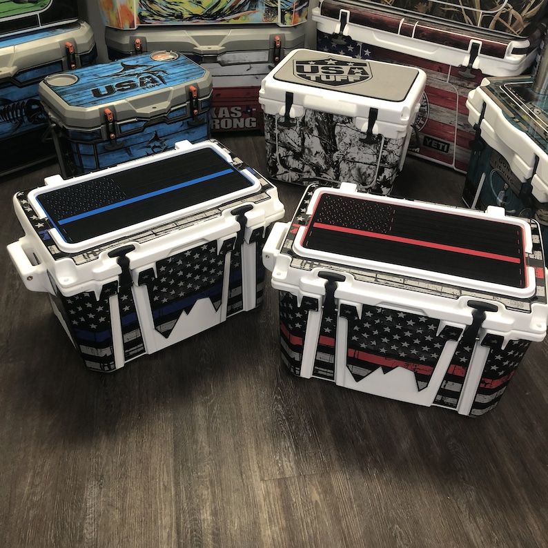 May include: Two white coolers with black and white American flag designs. One cooler has a blue stripe across the top and the other has a red stripe. Both coolers have the words "USA Tuff" on the top.